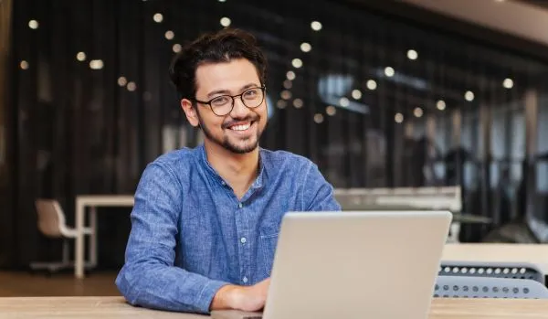 A happy young office worker is smiling at the camera over his laptop in an open working space.