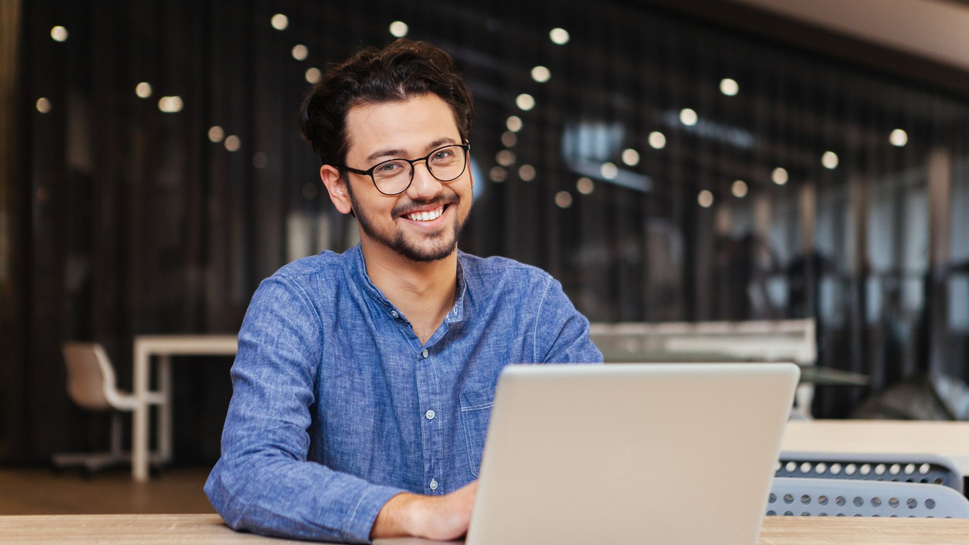 A happy young office worker is smiling at the camera over his laptop in an open working space.
