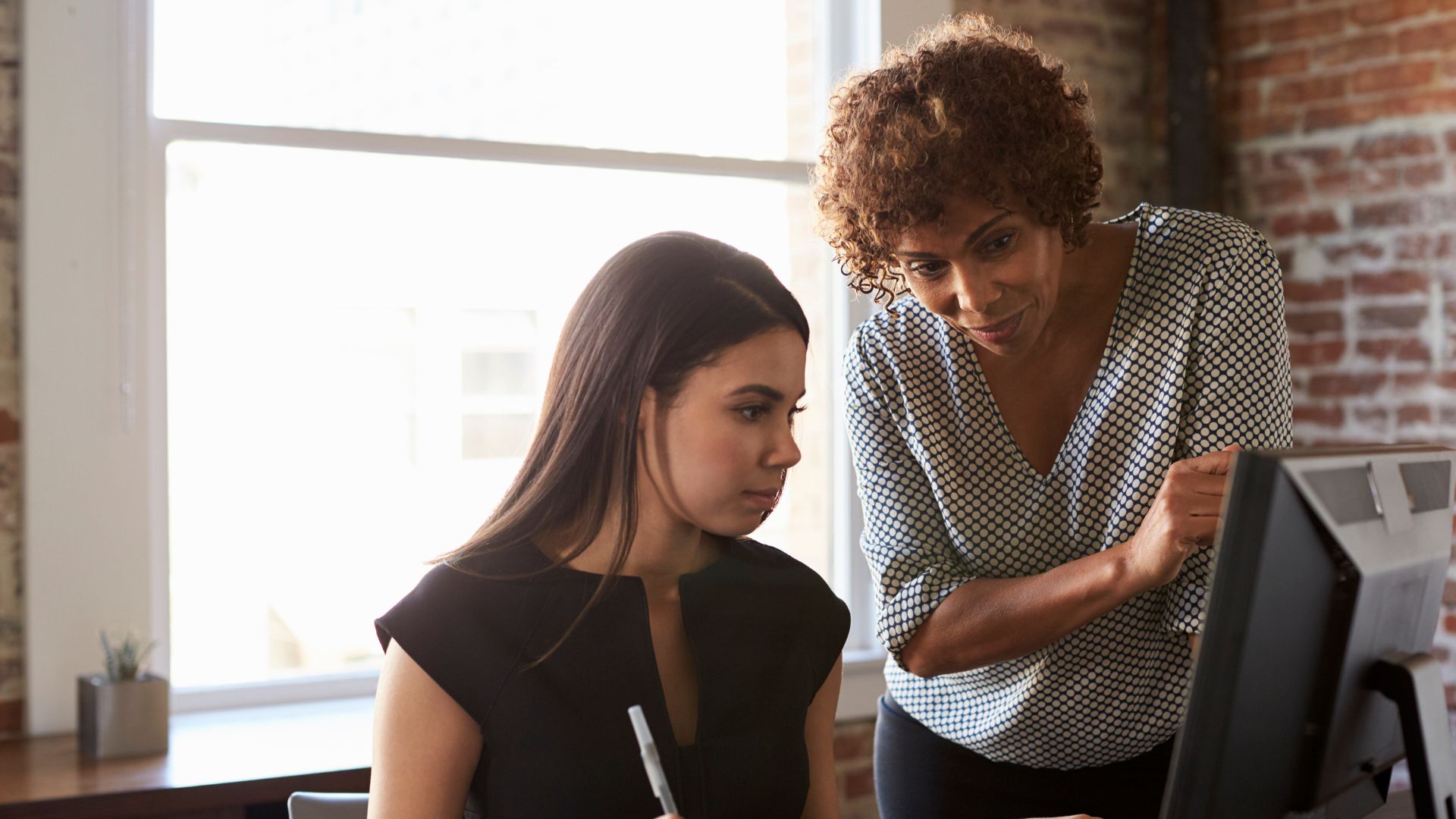 A woman is training a new manager at her desk. The new manager is sitting, while the woman stands beside her pointing at the monitor.