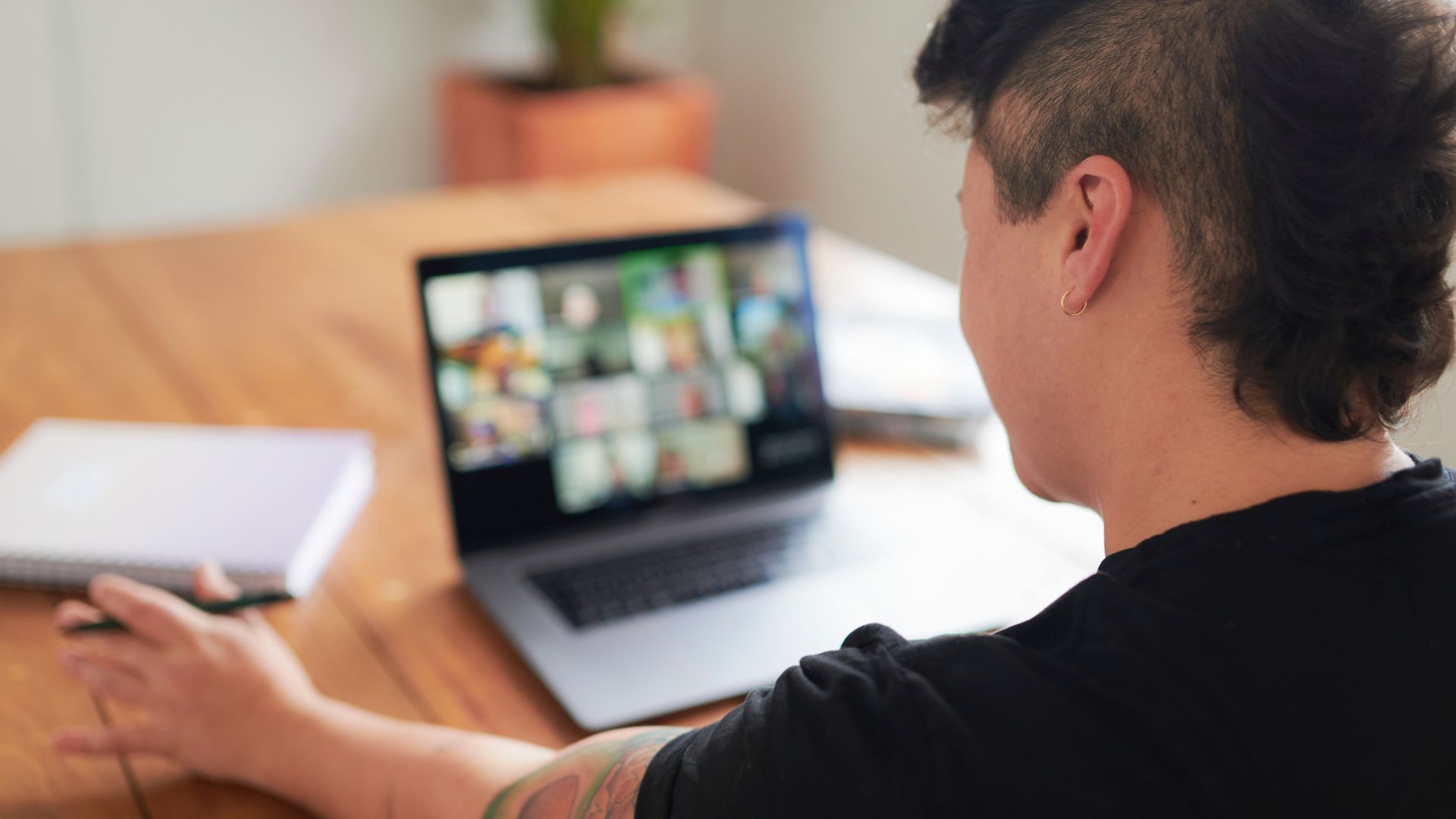 We're looking over the shoulder of a non-binary person at their laptop screen where they are participating in a live, virtual training course. 