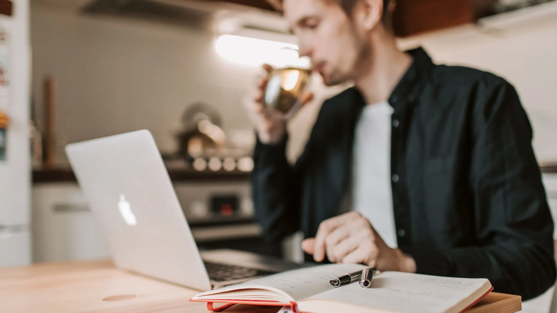 A man is taking a drink of water while working at his home desk with his laptop in front of him.