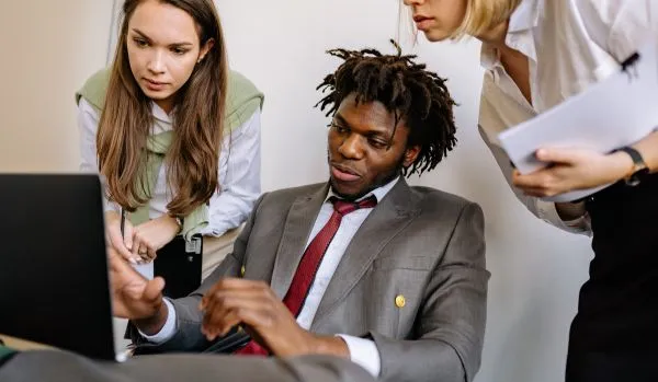 A manager is sitting at his computer, leading two of his team members through practical AI application. The two team members are standing behind him looking over his shoulder.