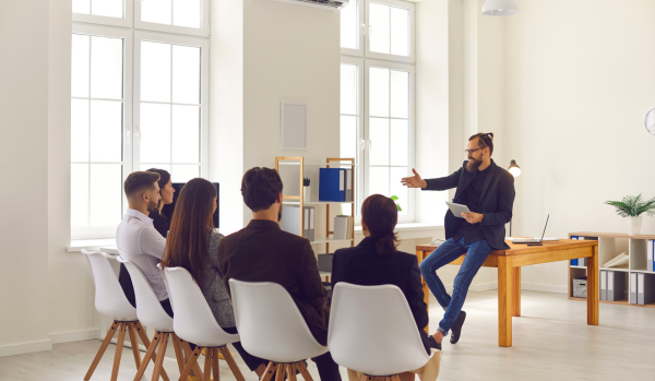 There are five people sitting in chairs in front of a man speaking who is leaning on a desk in an office setting.