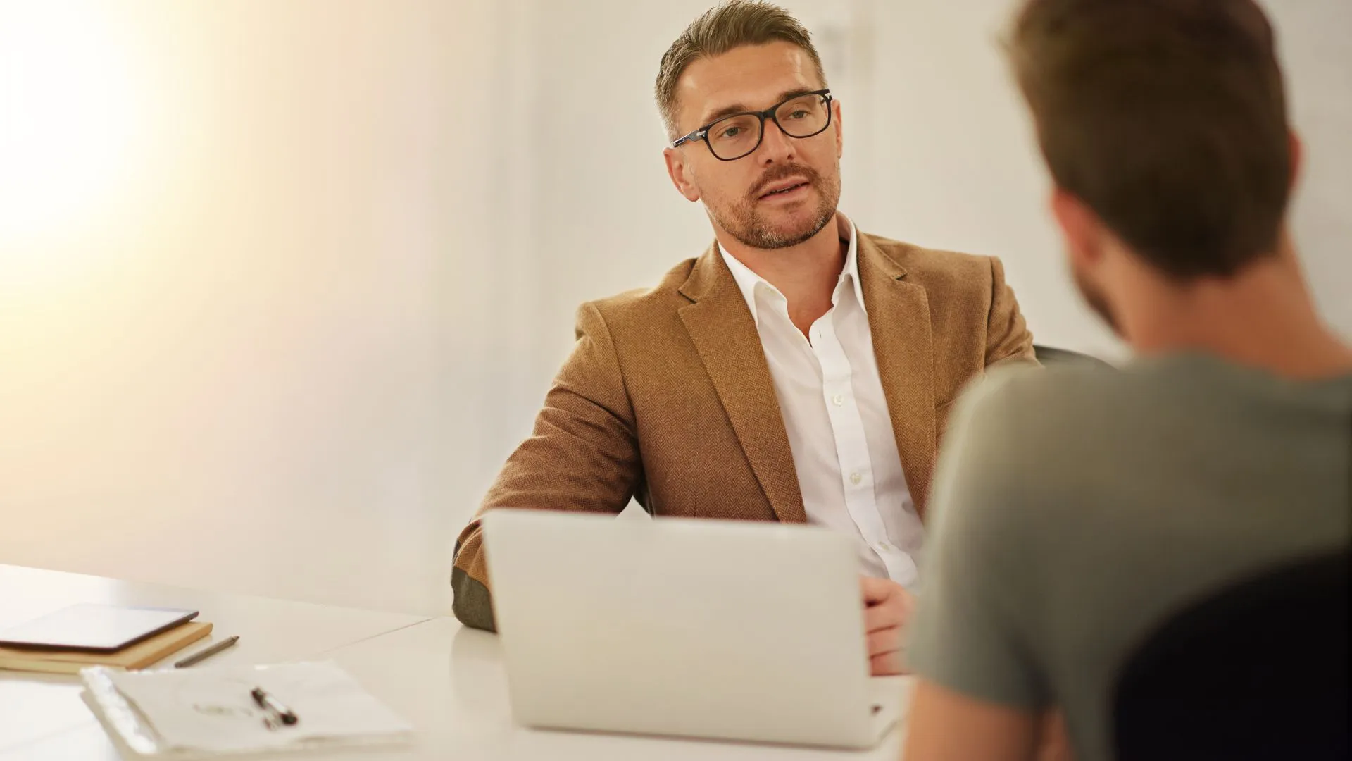 A man is having a feedback conversation with a member of his team. We are looking over the team member's shoulder at the man's face.