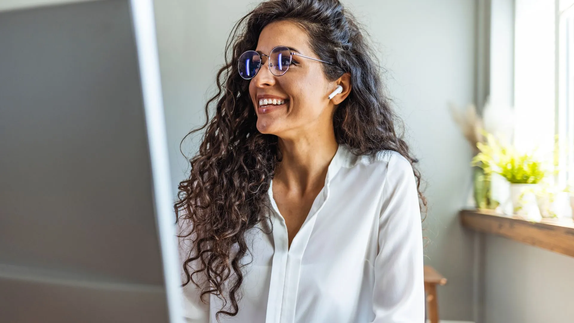 A woman is smiling at her monitor in her home office. She has her ear buds in.