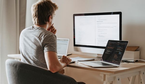 We're looking over a man's shoulder as he works on his laptop and monitor on his desk at home.