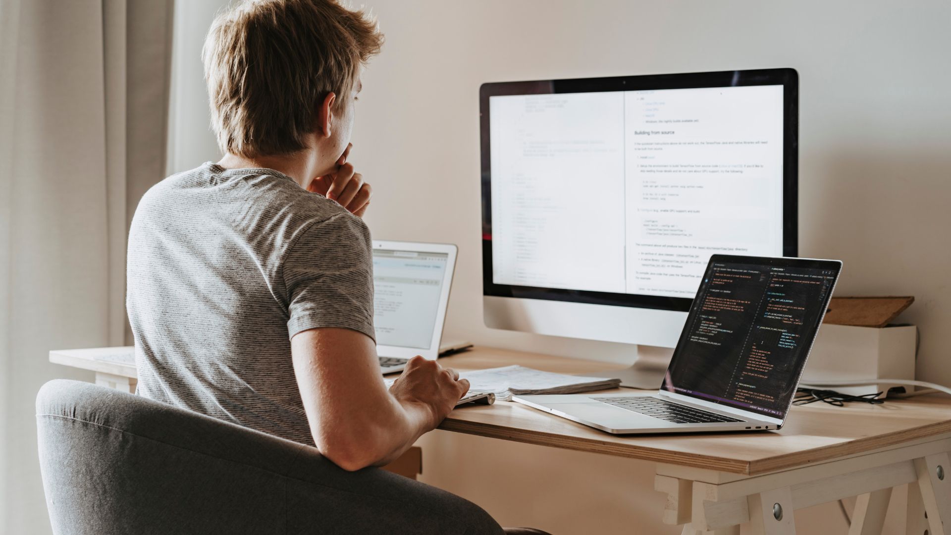 We're looking over a man's shoulder as he works on his laptop and monitor on his desk at home.