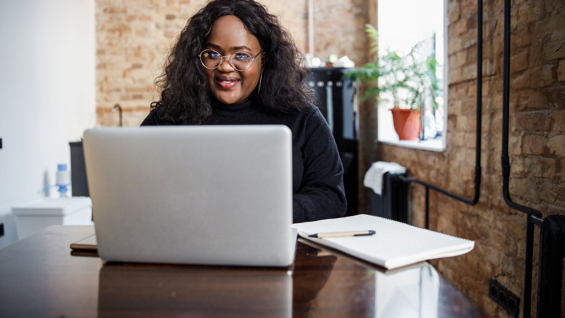 A woman is looking directly at the camera over the top of her laptop. She is wearing glasses and there is a notebook on the table next to her computer.
