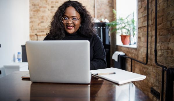 A woman is looking directly at the camera over the top of her laptop. She is wearing glasses and there is a notebook on the table next to her computer.