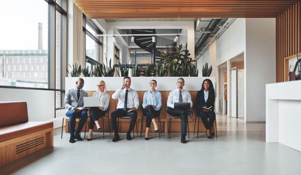 A diverse group of job applicants are sitting in a row in the lobby of a modern office. They are laughing and conversing.