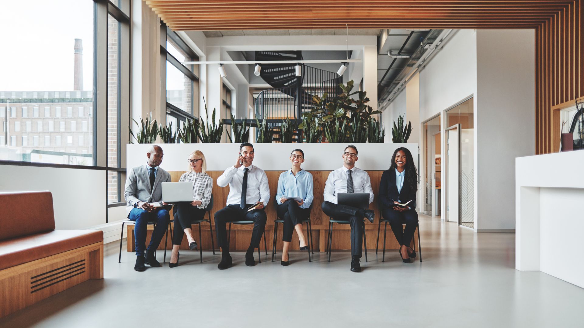 A diverse group of job applicants are sitting in a row in the lobby of a modern office. They are laughing and conversing.