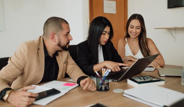 Three people are working together at a conference table. Two of them are looking over at the middle person's laptop.
