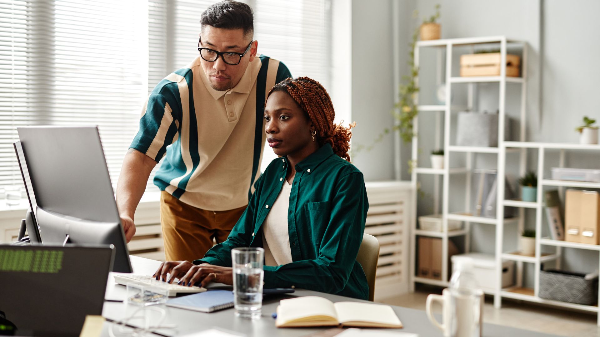 A man is standing behind a woman at a desk, pointing at something on her computer.