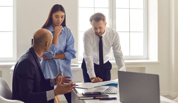 A boss is sitting at his desk having a conversation with two younger employees who are standing and showing him some documents.