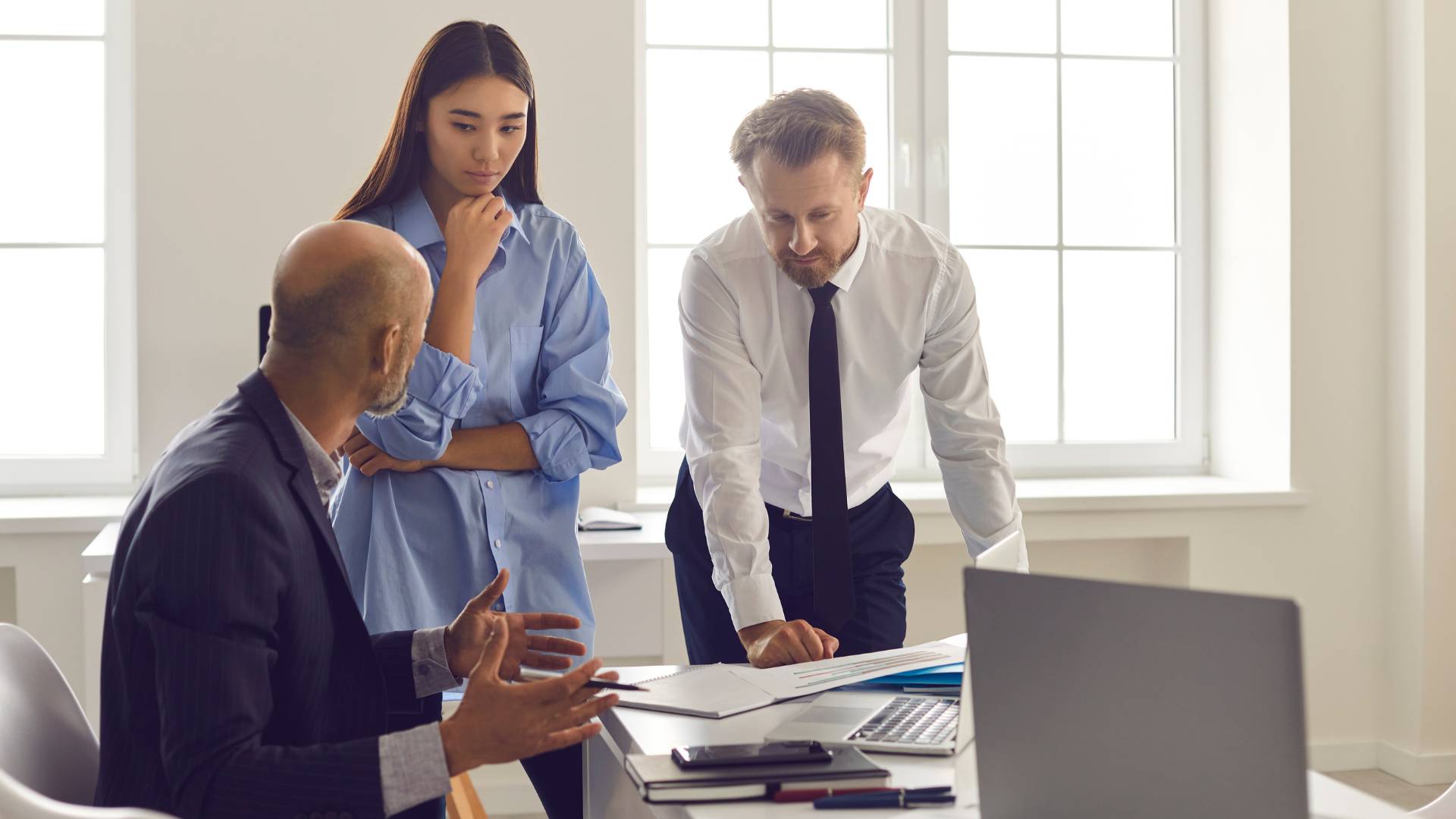 A boss is sitting at his desk having a conversation with two younger employees who are standing and showing him some documents.