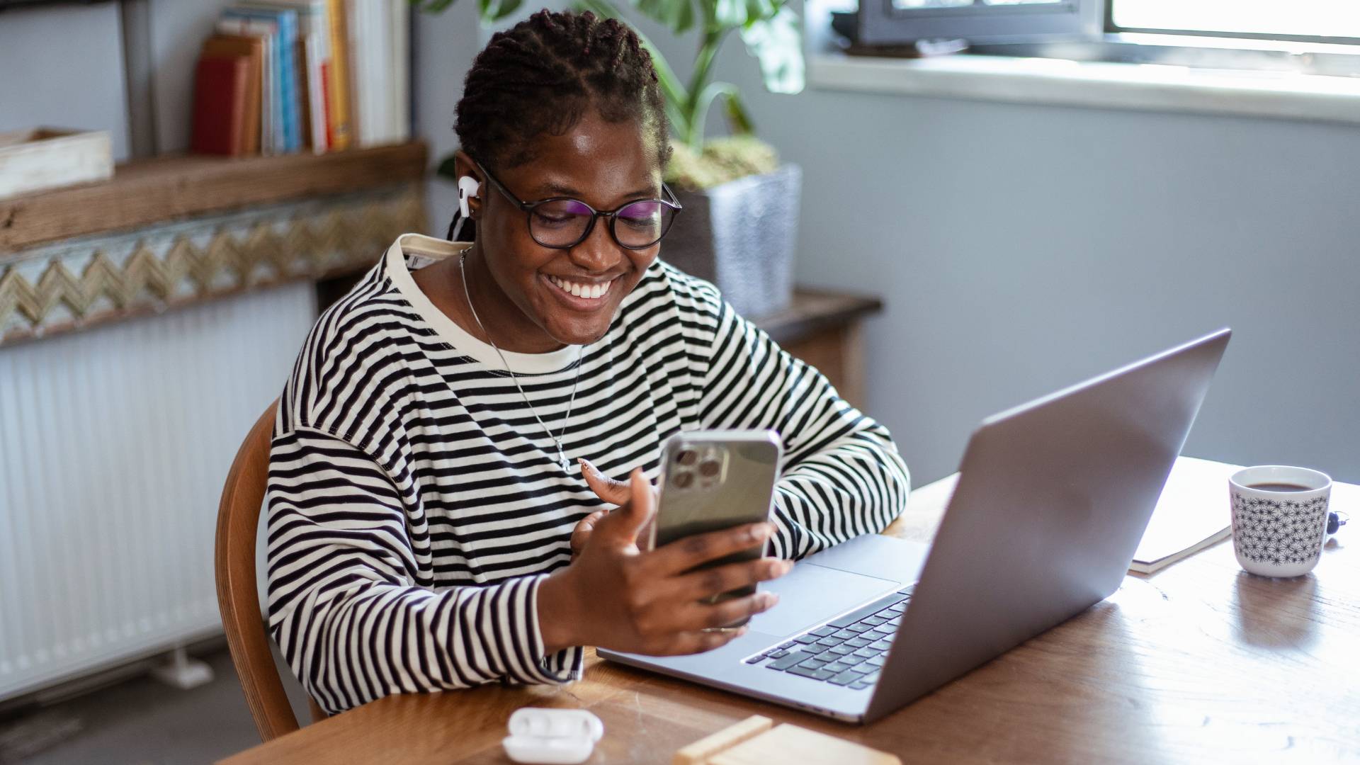 A young woman is working at her laptop on her desk, smiling and looking at her phone.