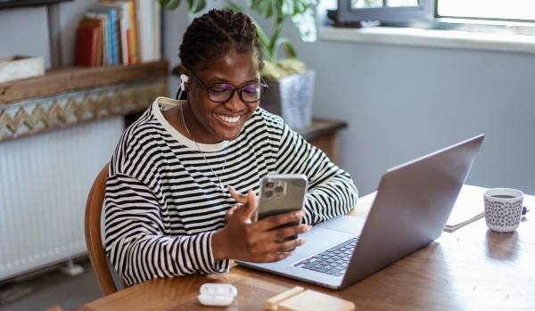 A young woman is working at her laptop on her desk, smiling and looking at her phone.