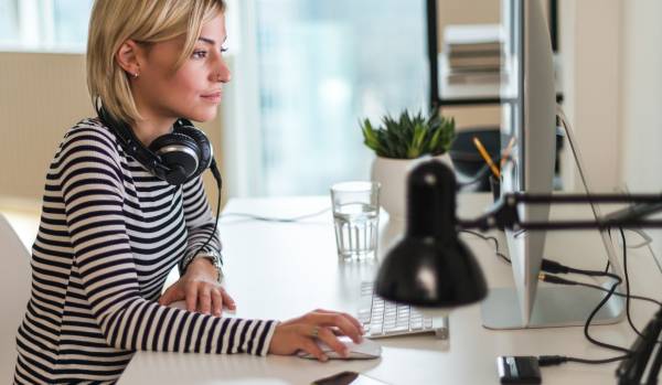 We are looking at the profile of a young woman working at her computer in her home. She has headphones around her neck.