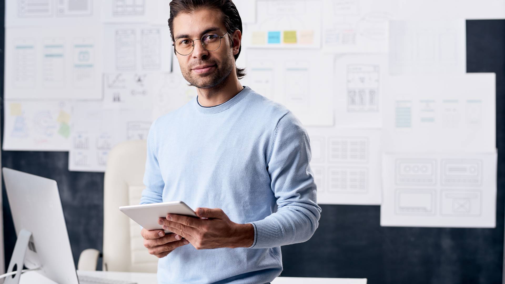 A project manager is standing in front of his desk and project flipcharts holding a tablet while looking into the camera with a slight smile.