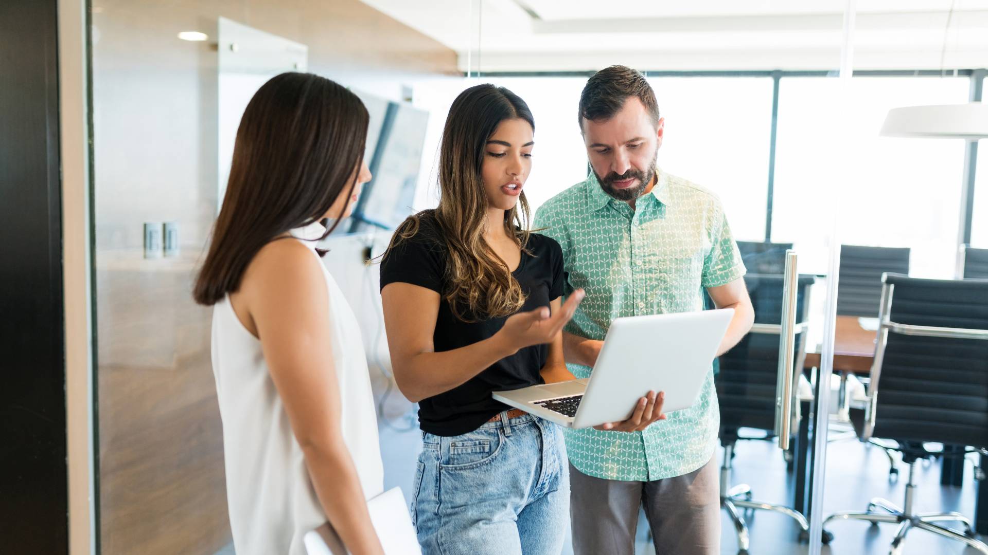Three people are standing speaking with each other in an office setting. The person in the middle is a woman and she's holding a laptop.