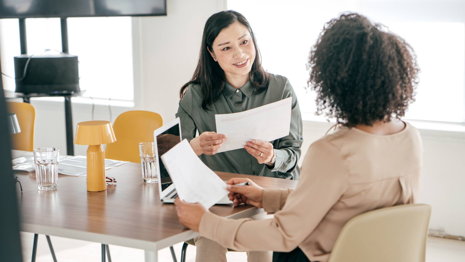 Two women are sitting at a table in an office talking. One of them is facing us while the other is facing away from us. They're going over documents they have in their hands and are sitting in front of a laptop.
