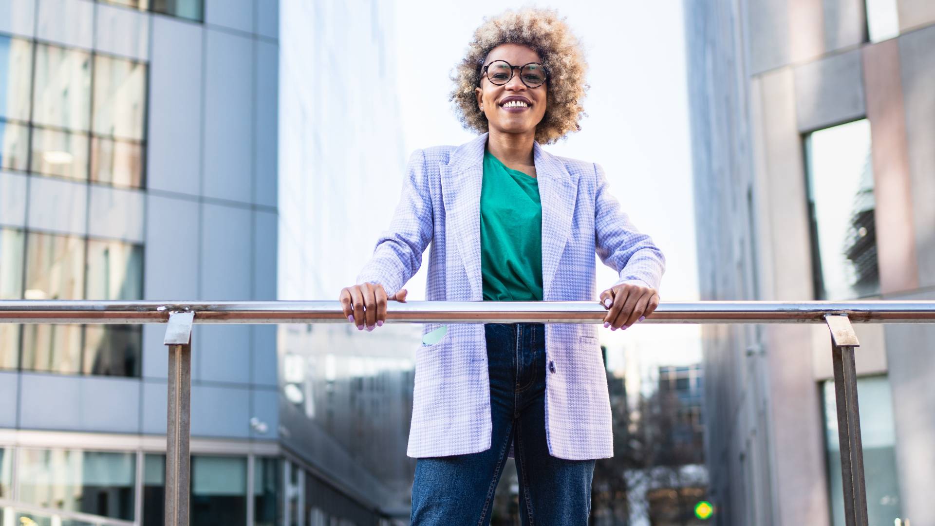 A smiling woman in glasses is looking at us from an elevated walkway, holding onto the railing.