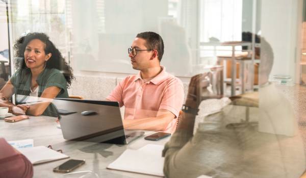 We are looking through a glass conference room wall at a diverse group of people participating in a training around a conference table.