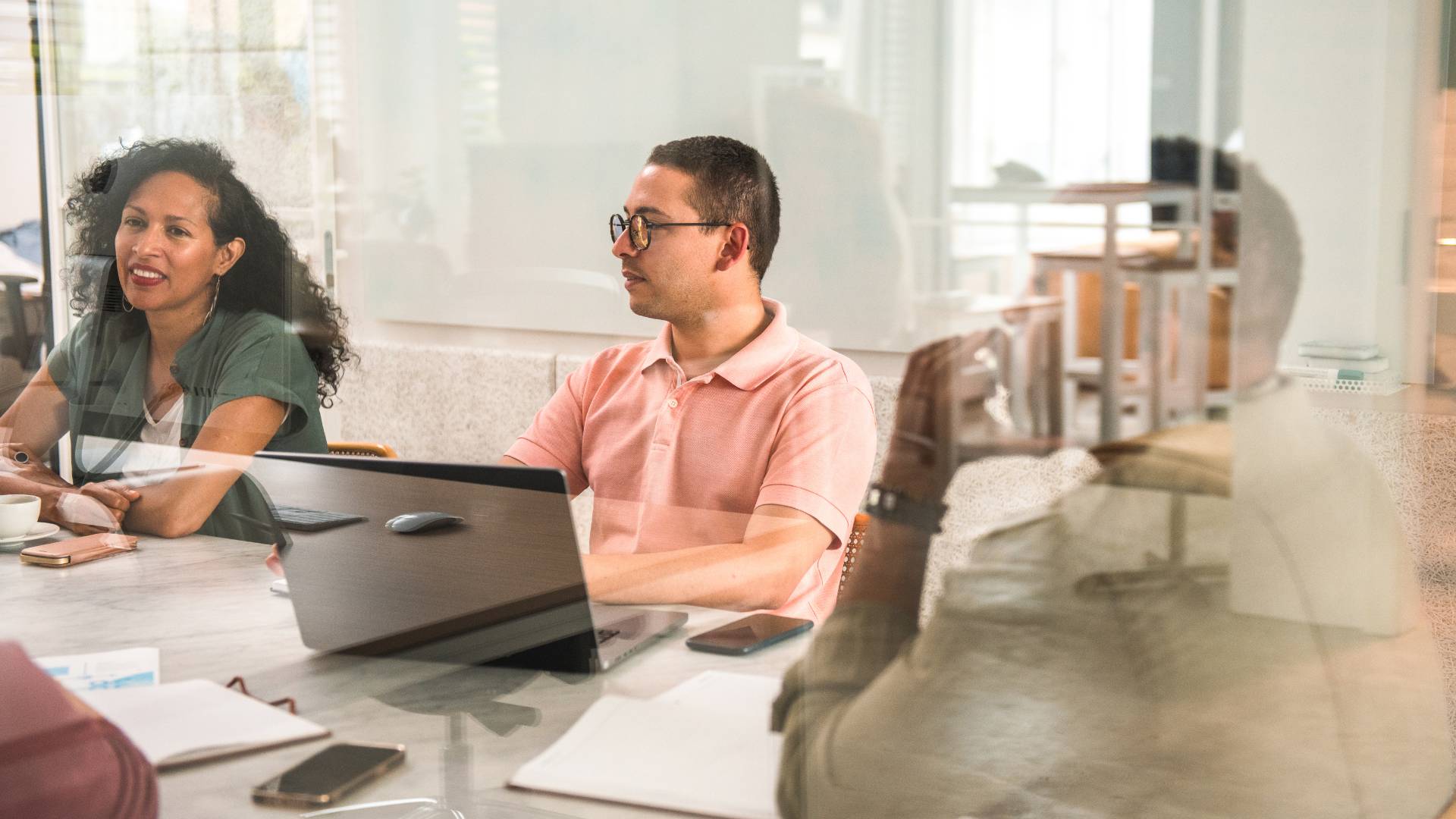 We are looking through a glass conference room wall at a diverse group of people participating in a training around a conference table.