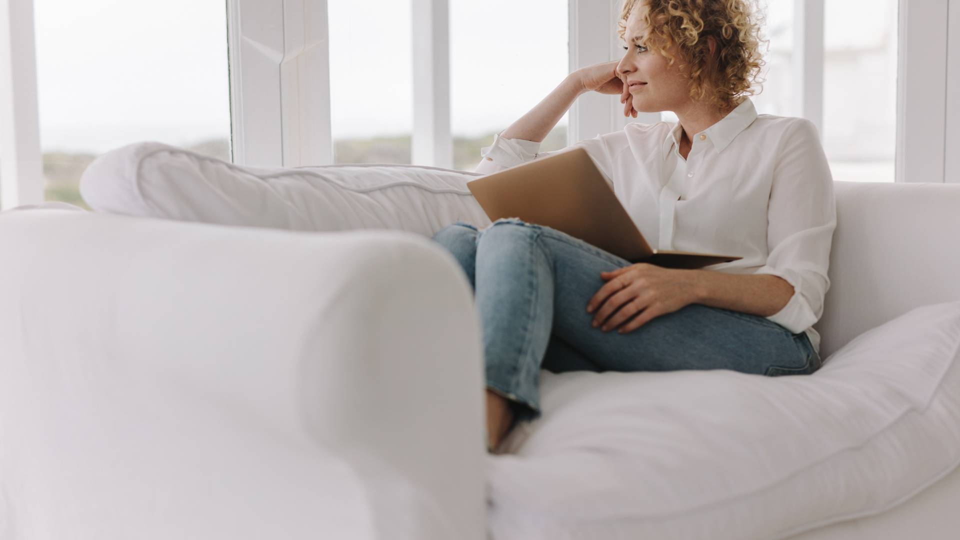 A woman is sitting on her couch, laptop in her lap, while looking out the window.