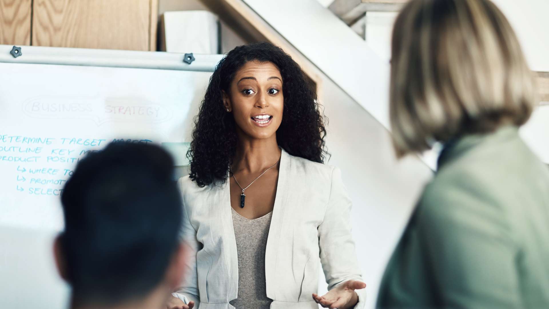 A young black woman is presenting to two colleagues. We are looking through the shoulders of the colleagues at her and her whiteboard.