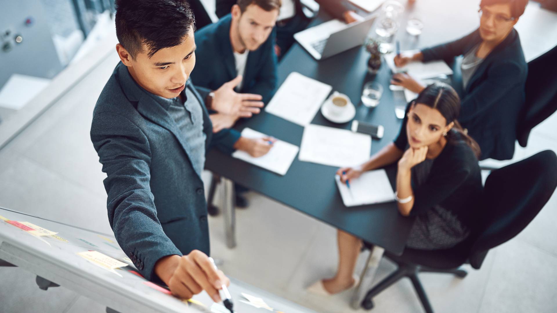 We are looking down at a man writing on a whiteboard while talking to other colleagues who are sitting around a conference table.