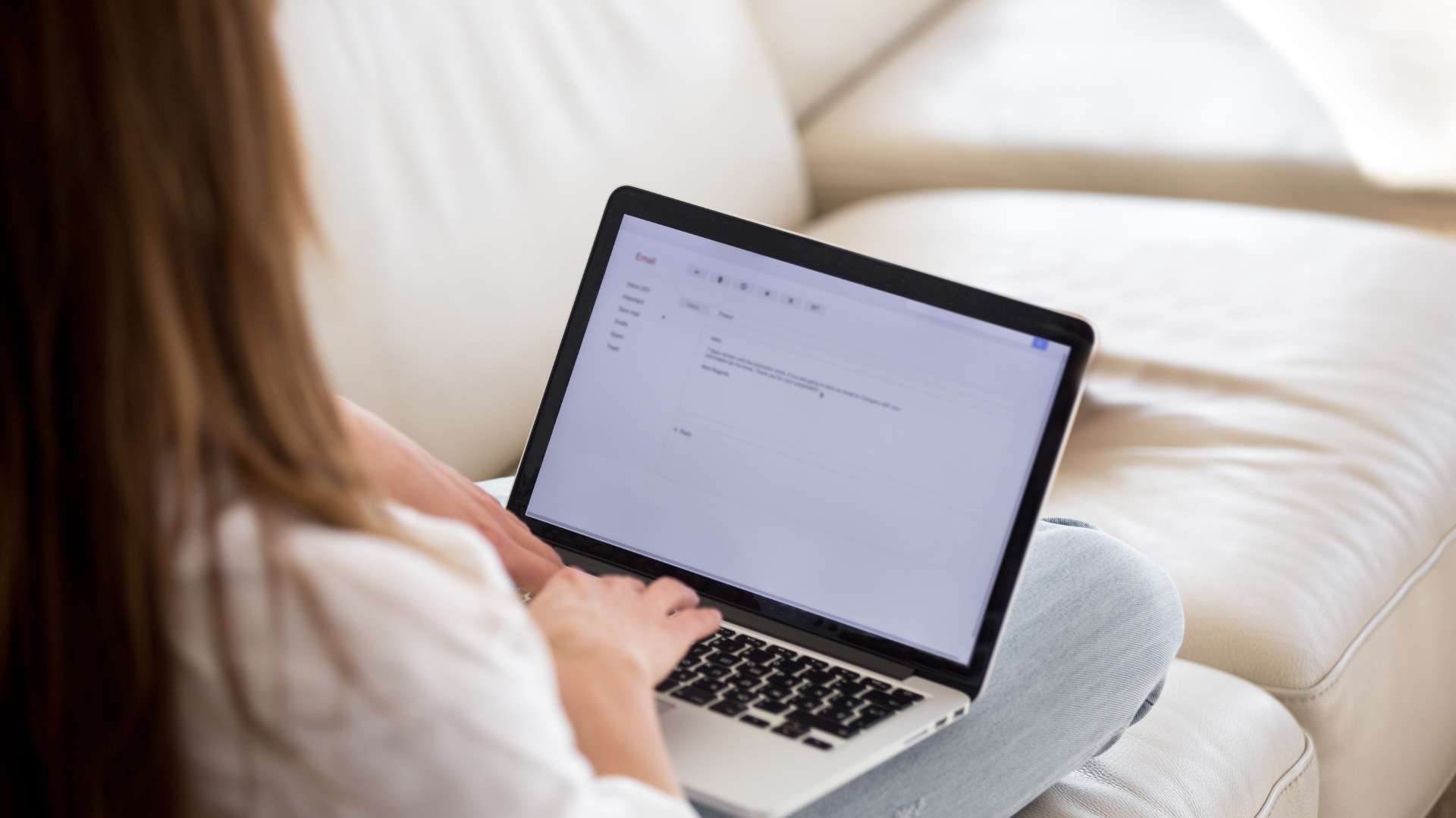 We're looking over a woman's shoulder as she composes an email on her laptop while working from her couch.