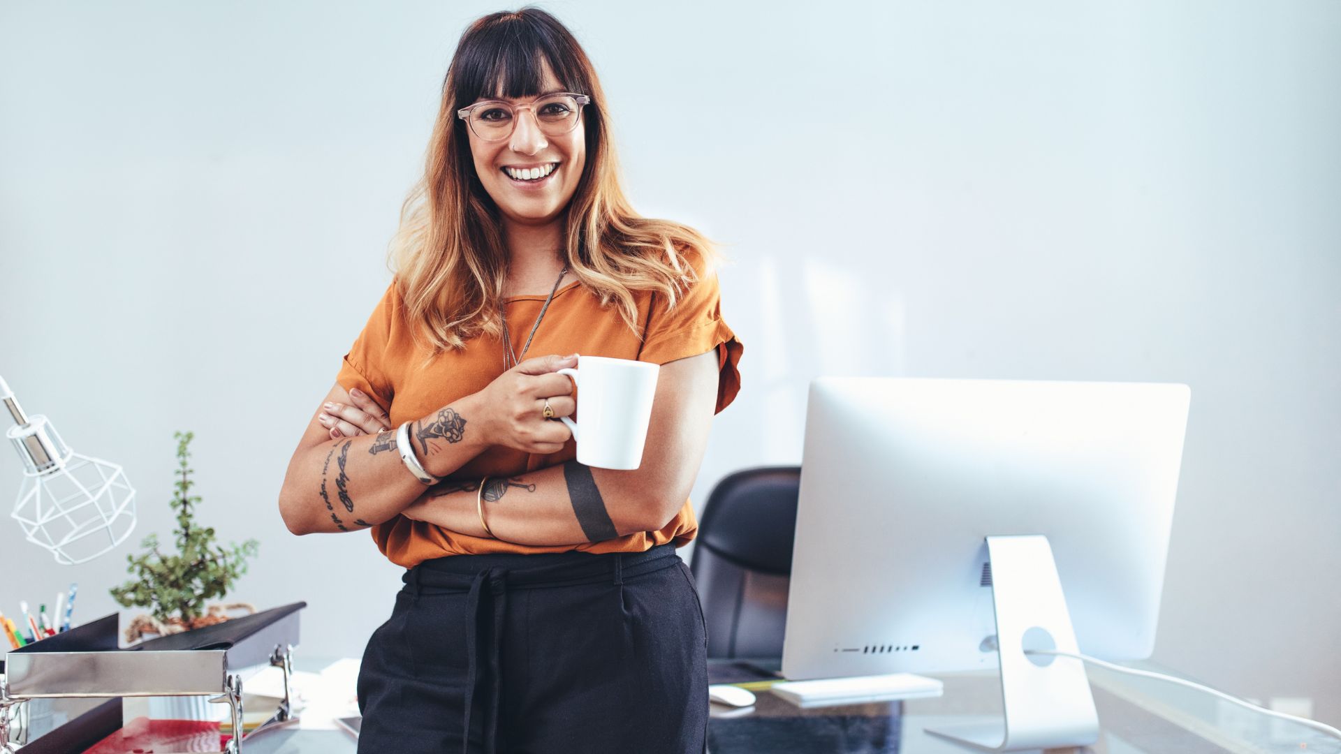 A young, tattooed female presenting manager is standing in front of her desk with her arms crossed holding a cup of coffee. 