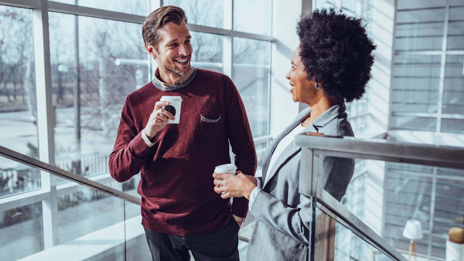 A man and woman are having a conversation on a stairwell in an office building. They're both holding coffee and smiling.
