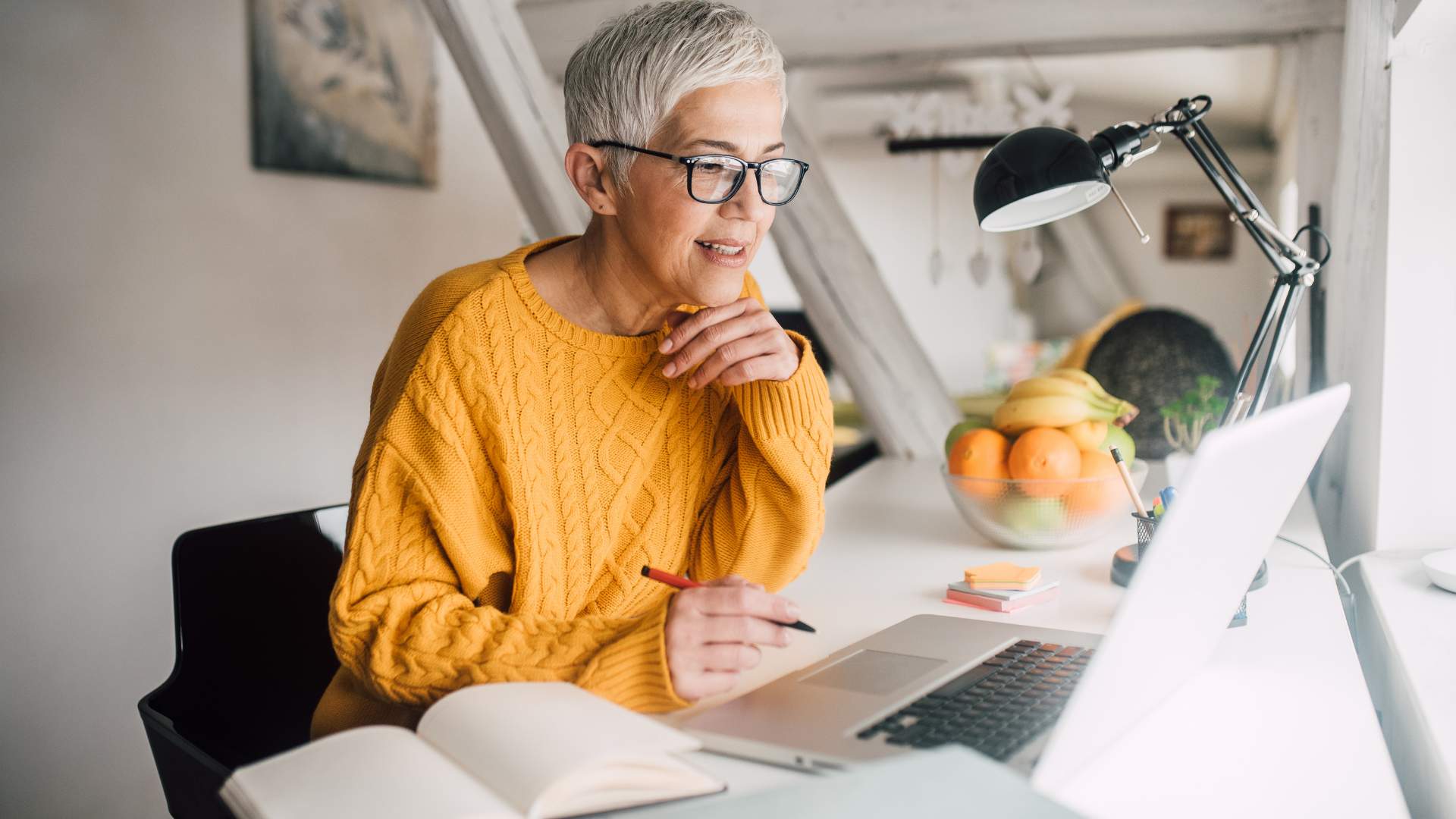 Older woman with a short, grey pixie cut in a yellow sweater is working on her laptop from home, ready to take notes in her notebook.