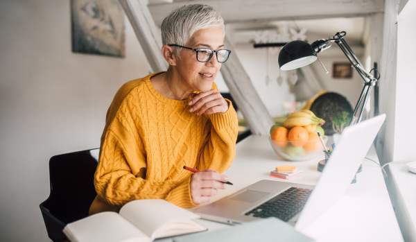 Older woman with a short, grey pixie cut in a yellow sweater is working on her laptop from home, ready to take notes in her notebook.