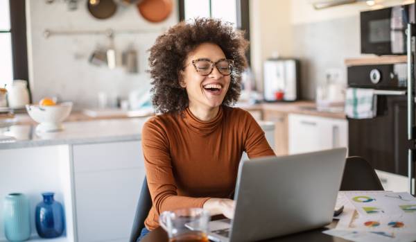 A woman in glasses is working from her kitchen table and smiling at her laptop.