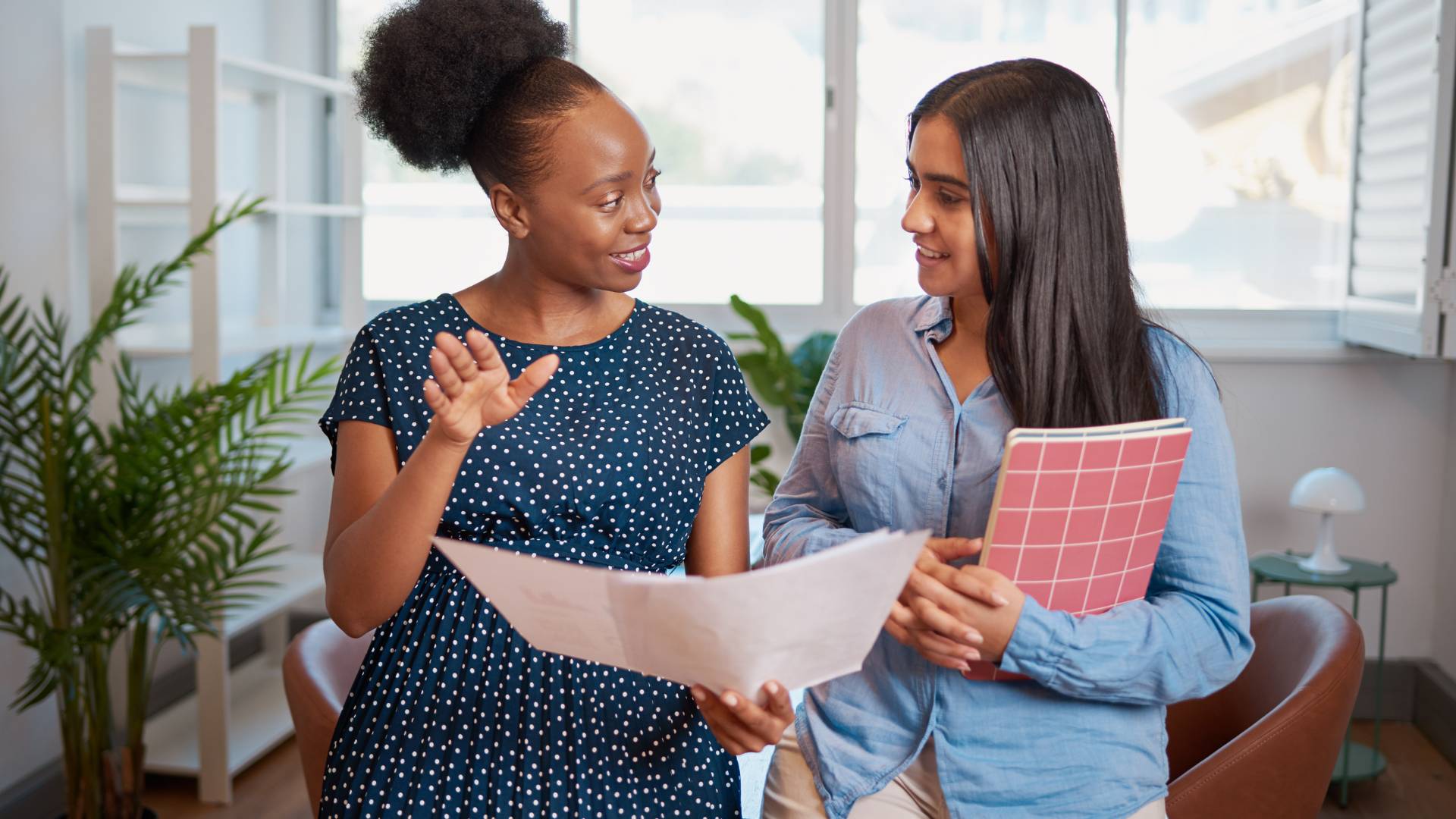 There are two women standing and talking in an office to each other. One woman is Black and the other woman is white.