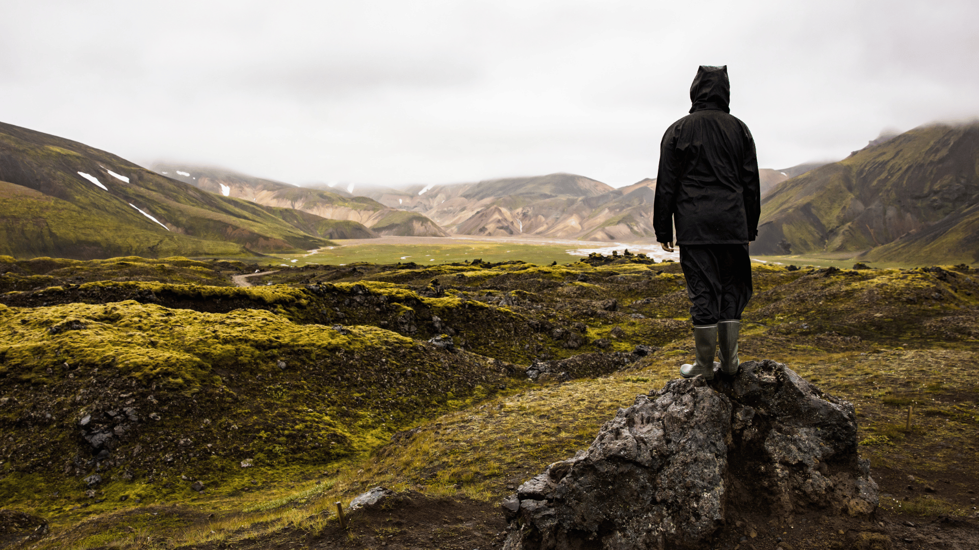 A person in rain gear standing on a rock with their back to us. They're looking out at a (possibly volcanic) valley with green moss on the surface.