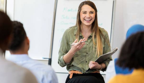 A woman is standing at the front of a meeting room leading a training. We are looking over the shoulders of the participants.