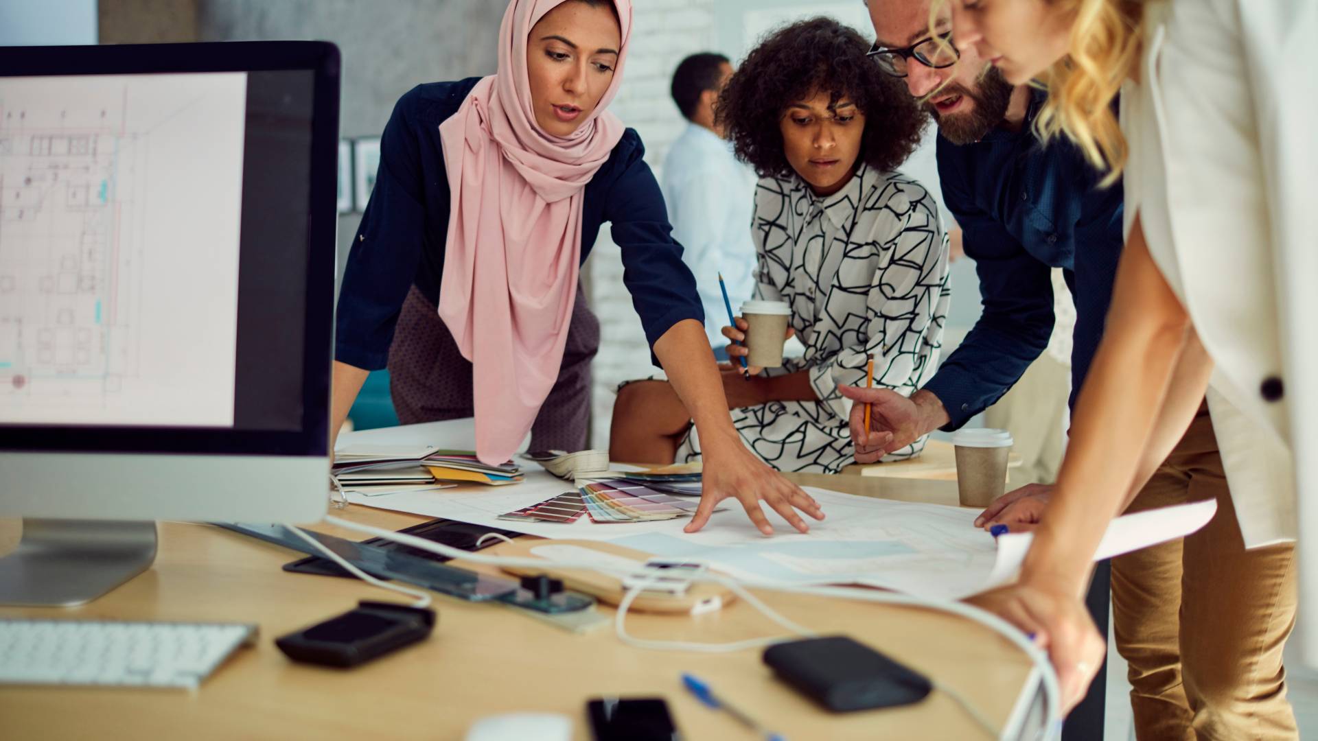 There are four employees having a meeting in a conference room standing gathered at a table looking down at documents. One employee is a woman and she's wearing a hijab.