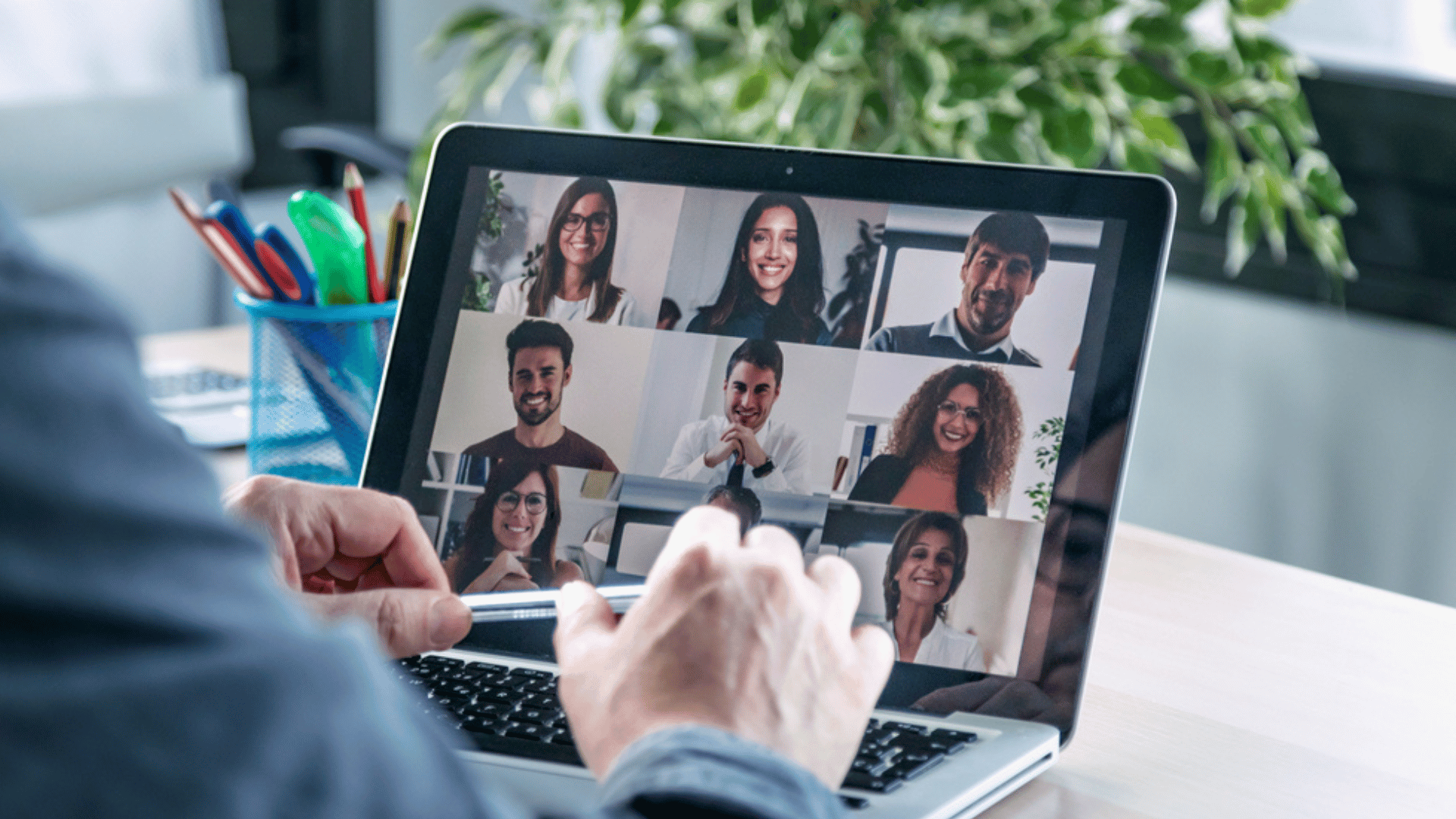 A person looking at his computer screen, filled with 9 Zoom boxes. We see only the person's right arm and two hands. The person is holding a pen with both hands.