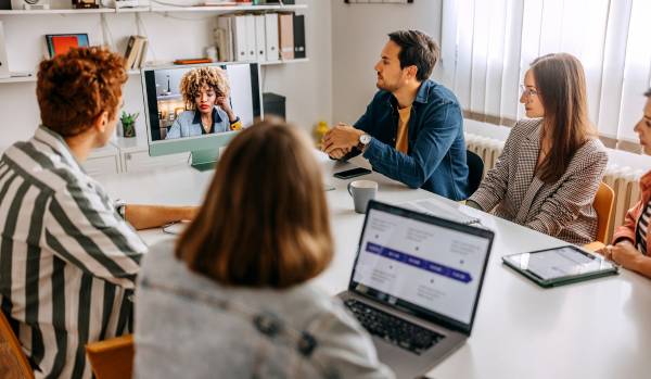 A diverse group of people sit around a conference table, listening to a colleague on a video conference.