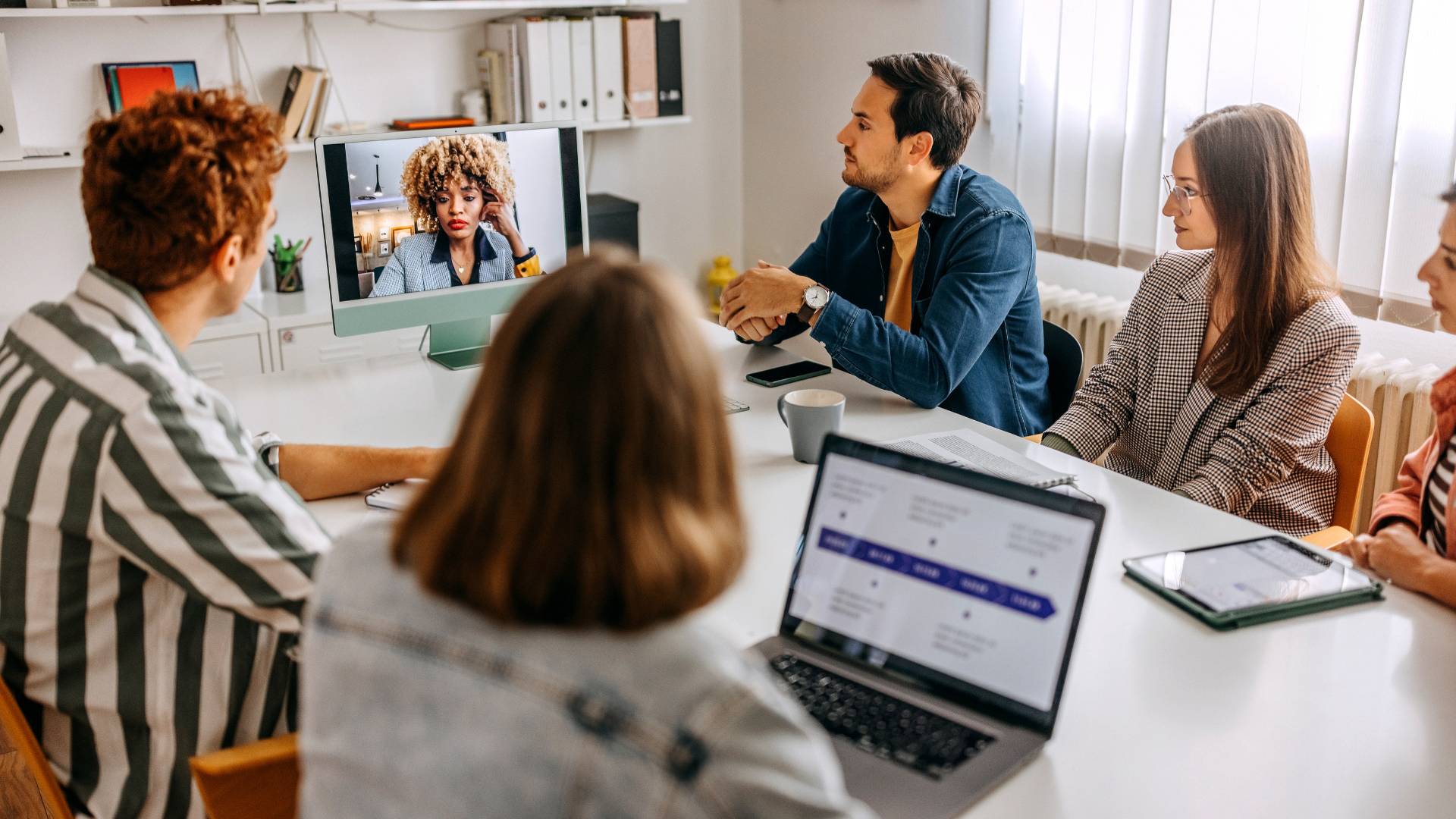 A diverse group of people sit around a conference table, listening to a colleague on a video conference.