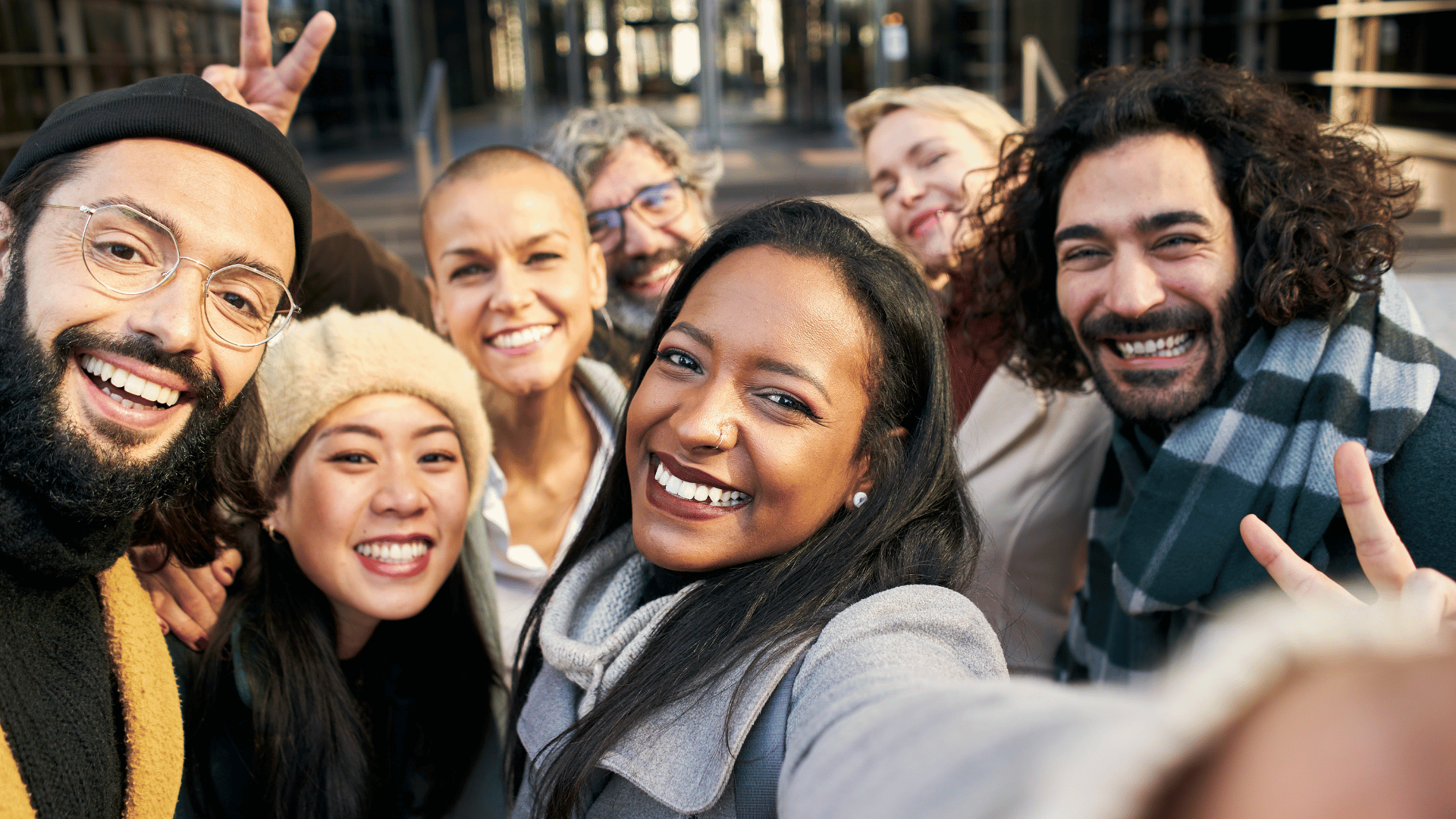 A team of diverse employees are standing outside an office building. They're smiling and one is holding out her arm to take a selfie. Someone in the back is holding up 2 fingers to make the peace sign.