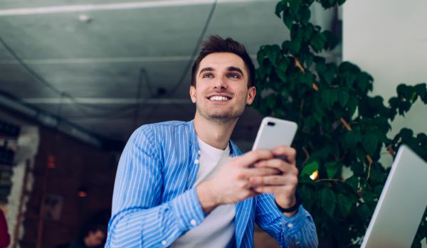 A young man is smiling off into the distance while holiday his phone in both hands.