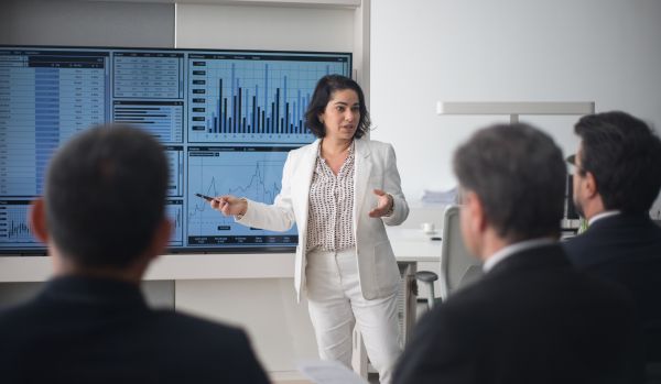 A woman in a white suit is presenting to a group of corporate executives. We are looking over the executives' shoulders.