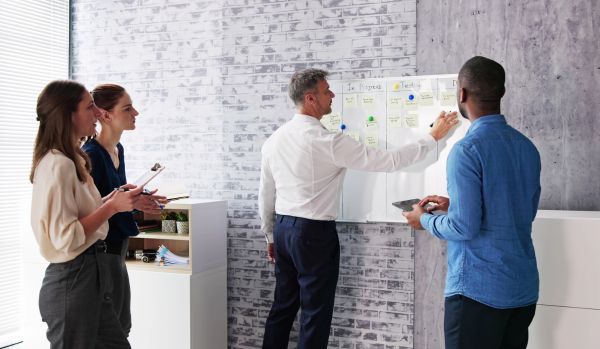 A diverse team of four people are looking at and working on a whiteboard, doing agile planning.