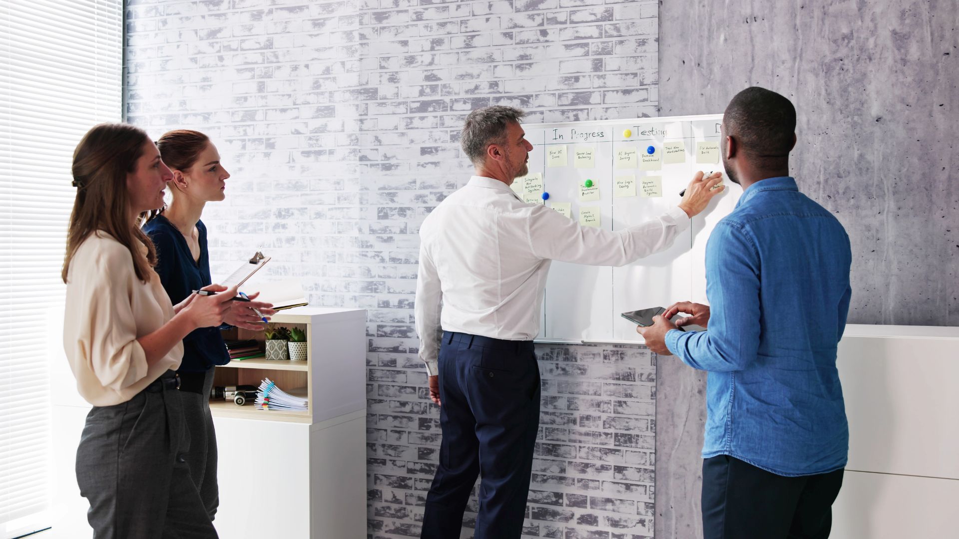 A diverse team of four people are looking at and working on a whiteboard, doing agile planning.