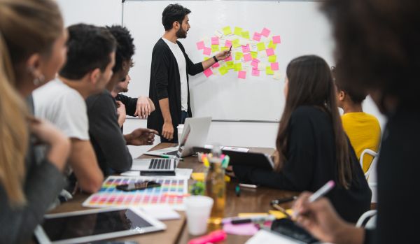 A diverse team of people are sitting around a conference table looking at their manager who is at the whiteboard discussing post-it notes that are stuck to it.